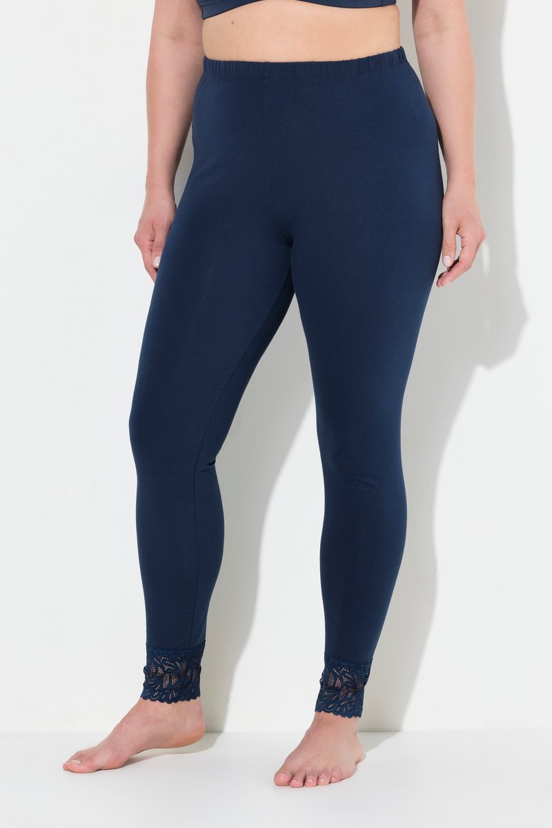Vrouw draagt marineblauwe legging met elastische taille en kanten randje bij de enkels, staande blootsvoets tegen een witte muur.