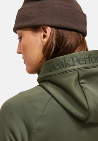 Moteris, vilkinti alyvuogių žalios spalvos bliuzoną su užrašu "Peak Performance" ant gaubto ir rudą megztą kepurę, žiūri į kairę prieš paprastą foną.