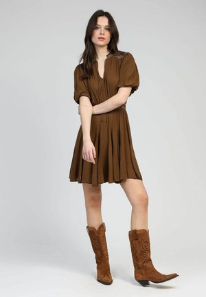 Robe marron à manches courtes avec une taille cintrée et une jupe évasée. Portée avec des bottes de cowboy en daim marron ornées de broderies délicates.