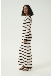 Robe maxi en tricot à rayures, noire et crème, avec des manches longues et une coupe ajustée. Accessoirisée avec des sandales à talons noires.