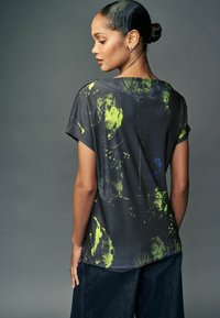 Femeie cu părul închis la culoare prins într-un coc, purtând un tricou negru cu modele abstracte în galben neon și albastru, stând lateral pe un fundal gri.