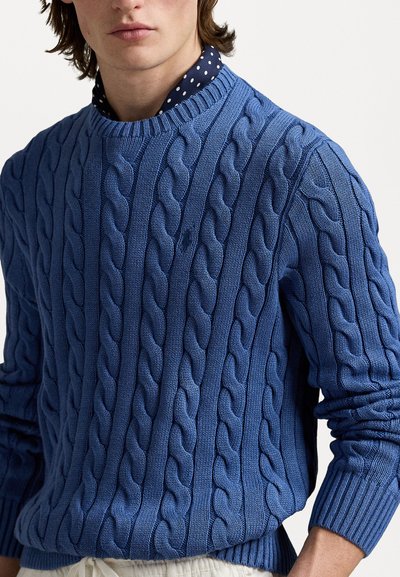 Blå kabelstrikket sweater med ribbet halsudskæring og manchetter. Har et struktureret mønster af sammenflettede kabler. Bæres over en polkadot skjorte med krave.