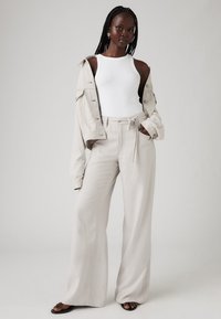 Žena s dugim pletenicama, odjevena u bijelu majicu bez rukava, bež široke hlače, usklađenu jaknu spuštenu s ramena i crne sandale, stoji.