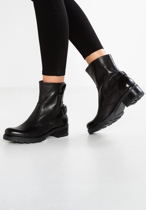 Person trägt schwarze Leder-Stiefeletten mit seitlichem Reißverschluss und Schnallendetail, kombiniert mit eng anliegenden schwarzen Hosen vor weißem Hintergrund.