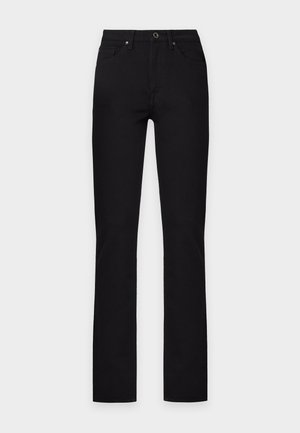 Une femme porte une surchemise beige, un col roulé blanc, un pantalon noir et des chaussures à talons marron sur un fond clair uni.