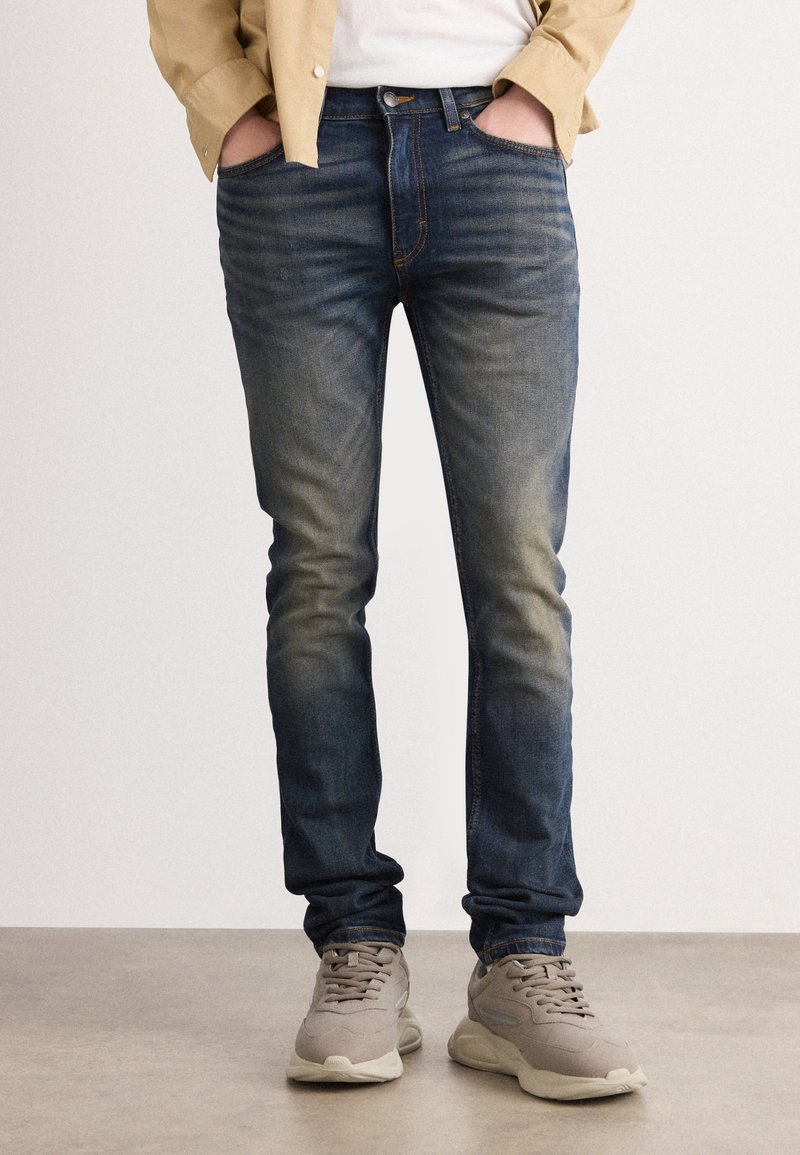 Homme portant un jean slim bleu délavé avec les mains dans les poches et des baskets beiges, debout sur un sol lisse.