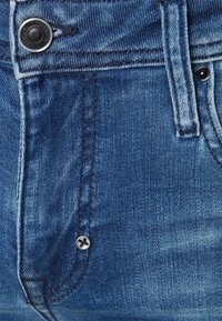 Modré džínové kalhoty s detailním pohledem na kovový knoflík, kontrastní stehy a hladkou texturu. Obsahují kapsy a poutka na opasek.