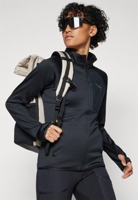 Black zippered jacket with a textured finish, paired with a gray and black striped strap backpack. Features round sunglasses and athletic leggings.