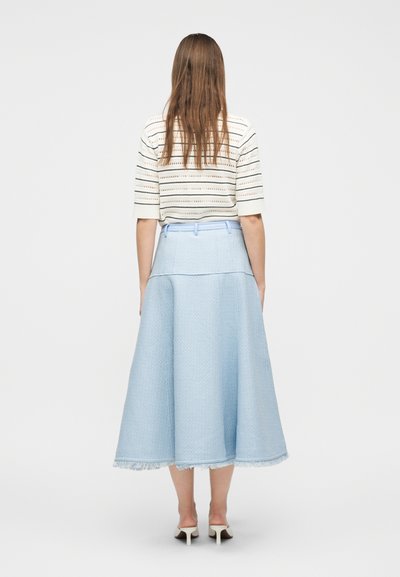 Γυναίκα που στέκεται προς τα πίσω φορώντας ριγέ κοντομάνικη μπλούζα, φούστα μέτριας μήκους ανοιχτού μπλε χρώματος με ξεφτισμένο τελείωμα και λευκές σανδάλες με τακούνι.