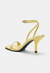 Gelbe High Heels mit quadratischem Zehendesign, ausgestattet mit flachen Knöchelriemen und einer schlanken schwarzen Sohle. Glattes Material mit minimalen Akzenten.
