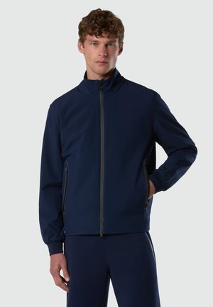 Giovane uomo con capelli ricci che indossa una giacca blu navy con zip e pantaloni coordinati, in piedi con una mano in tasca.