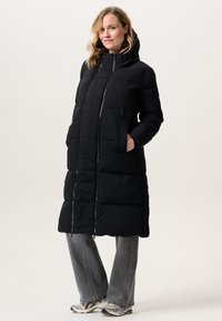 Vrouw in een lange zwarte pufferjas met capuchon, grijze spijkerbroek en witte sneakers, staand met handen in de zakken, licht glimlachend.