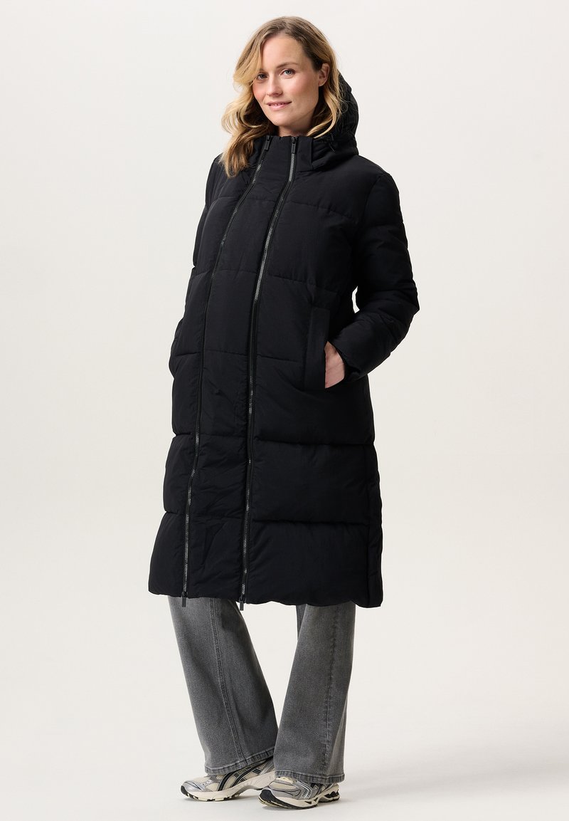 Vrouw in een lange zwarte pufferjas met capuchon, grijze spijkerbroek en witte sneakers, staand met handen in de zakken, licht glimlachend.