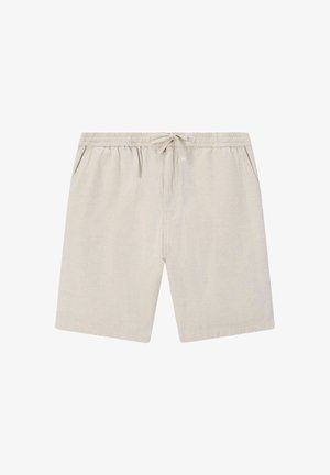 Homme portant un short beige, un t-shirt mauve et un sweat-shirt posé sur les épaules, debout, une main dans la poche, regardant devant.