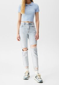 Polo cropped azul claro, jeans de lavado claro desgastados con roturas en las rodillas y zapatillas grises con acentos en crema. Diseño sencillo y ceñido.