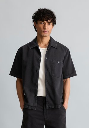 Junger Mann mit lockigem Haar trägt ein schwarzes kurzärmeliges Hemd über einem weißen Shirt, Hände in den Taschen, neutraler grauer Hintergrund.