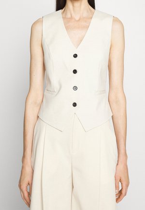 Femme portant un gilet crème sans manches avec quatre boutons noirs, associé à un pantalon crème plissé taille haute assorti.