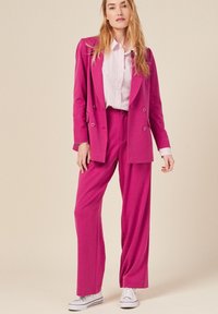 Fuchsia dubbelzijdig colbert met wijde broek en een gestreepte witte shirt eronder. Geaccentueerd met knopen en gedragen met witte sneakers.