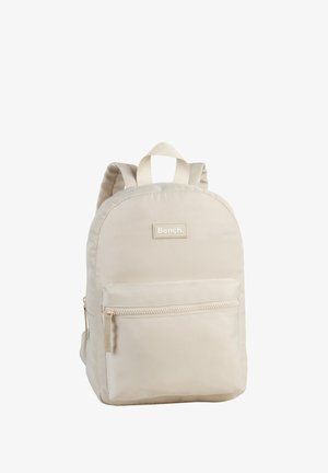 Light beige fabric backpack with rounded shape, a front zip pocket, two shoulder straps, and a top loop. Brand logo displayed on the front.