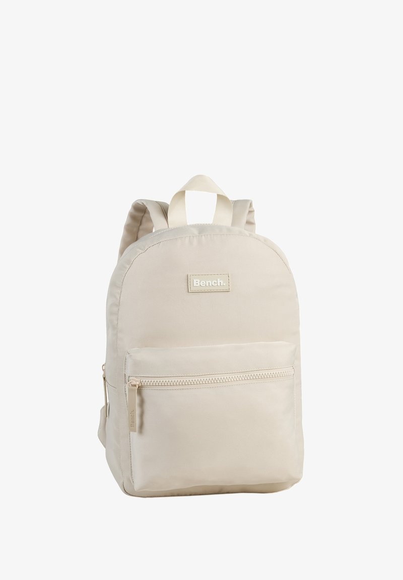 Hellbeige Stoff-Rucksack mit runder Form, einer frontalen Reißverschlusstasche, zwei Schulterträgern und einem oberen Griff. Markenlogo auf der Vorderseite abgebildet.