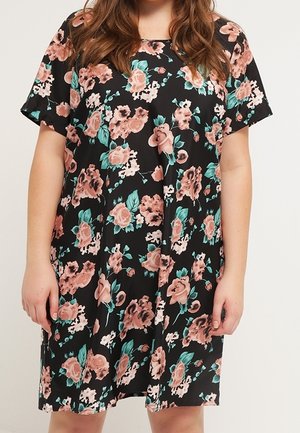 Frau trägt ein schwarzes, kurzärmeliges Kleid mit einem pink-grünen Blumenmuster und steht vor einem einfarbigen hellen Hintergrund.