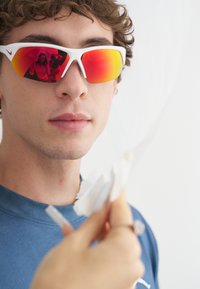 Άσπρα αθλητικά γυαλιά ηλίου με κόκκινους-πορτοκαλί φακούς που καθρεφτίζουν. Μαγαρισμένα μαλλιά και μπλε πουκάμισο στο προσκήνιο, θολό χέρι που κρατάει ένα πανί.