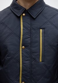 Μπλε μπουφάν με επένδυση από κουβέρτα, με μυτερό γιακά, κίτρινα στοιχεία κατά μήκος της λωρίδας των κουμπιών και τσέπη στο στήθος με αντίστοιχη διακόσμηση.