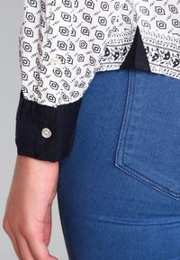 Camicia bianca con un motivo geometrico nero, polsini blu navy con bottoni; abbinata a jeans blu, evidenziando un design aderente e un tessuto testurizzato.