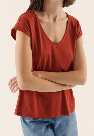 Personne aux cheveux courts et bruns portant un t-shirt à col en V couleur rouille et un jean bleu, les bras croisés sur la poitrine.