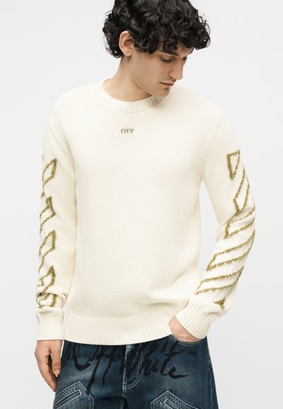 Νεαρός με κατσαρά μαλλιά φοράει λευκό πλεκτό πουλόβερ με χρυσά γεωμετρικά σχέδια στα μανίκια και το κείμενο "OFF" στο στήθος, συνδυασμένο με μπλε τζιν.