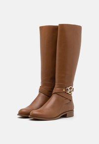 Bottes montantes en cuir marron avec un accent de logo doré et un détail de sangle. Texture lisse, bout arrondi et design à talon bloc bas.