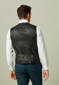 Mann in einem schwarzen Anzugs-Westen mit einstellbarem Riemen über einem weißen Hemd und blauen Hosen, der mit dem Rücken zur Kamera steht.