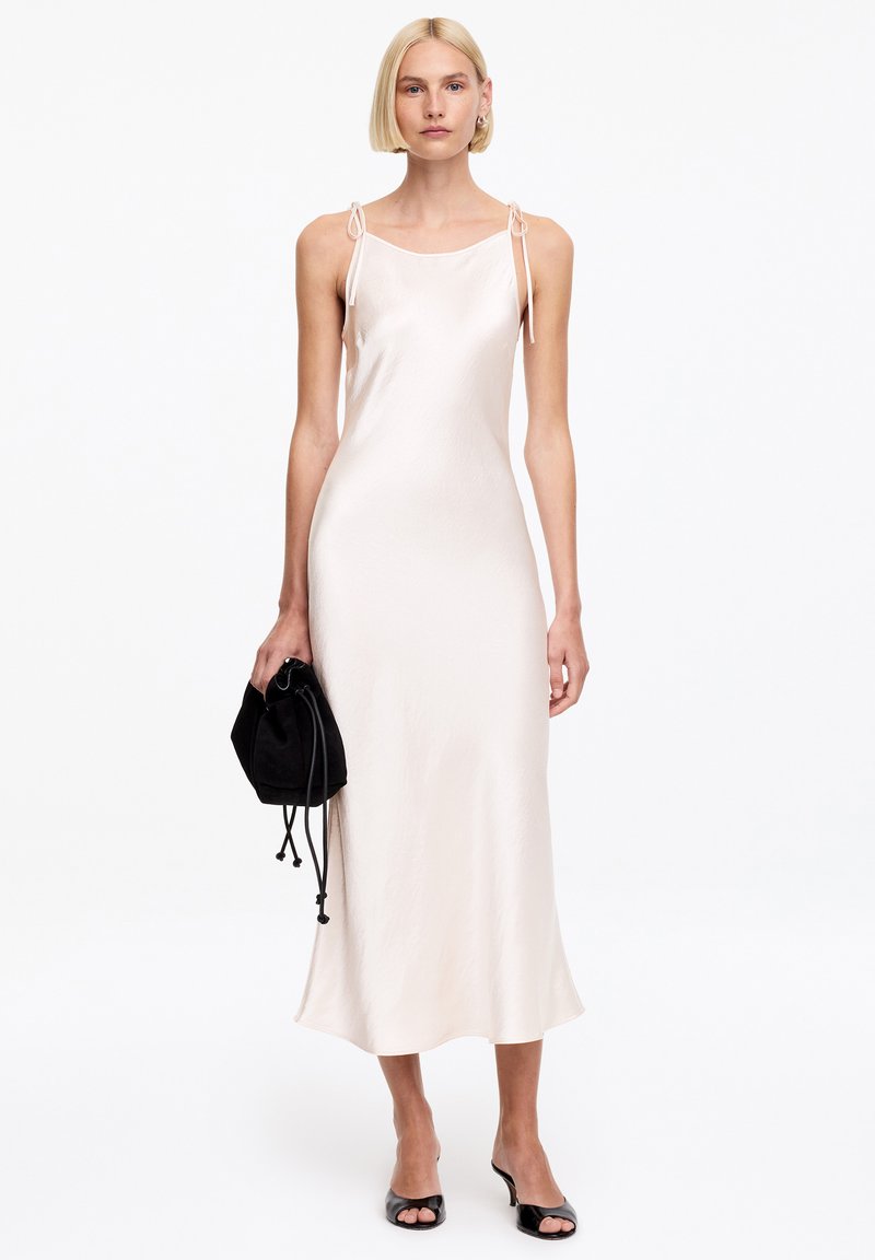 Femme aux cheveux blonds courts portant une robe midi rose clair sans manches avec des bretelles nouées aux épaules, tenant un sac à cordon noir et des talons noirs.