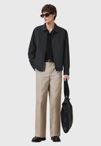 Zwarte zip-up jas, geribde zwarte trui, beige broek, zwarte schoenen, houdt een zwarte tas vast, draagt zwarte zonnebril; casual look.