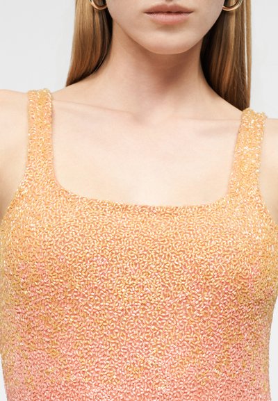Νεαρή γυναίκα που φοράει αμάνικο τοπ πυκνά καλυμμένο με μικρές ροδακινί και χρυσές χάντρες, με εμφανές το ντεκολτέ και χρυσούς κρίκους σκουλαρίκια.