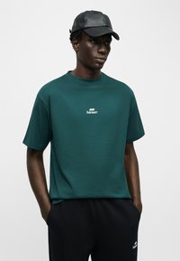 Άνδρας που φοράει σκούρο πράσινο t-shirt Bareen και μαύρο παντελόνι, με τα χέρια στις τσέπες, φορώντας μαύρο δερμάτινο καπέλο, σε απλό γκρι φόντο.