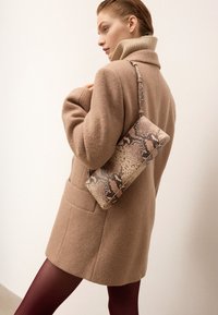 Donna con cappotto di lana beige e collant bordeaux, che tiene una borsa con motivo rettile sulla spalla, guardando di lato.