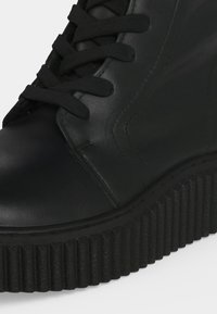 Botte à plateforme en cuir noir avec semelle épaisse rainurée et lacets noirs, montrant un gros plan des coutures et de la partie supérieure avant.