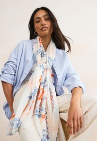 Hellblaues Hemd zum Knöpfen, cremefarbener Schal mit blauen und orangefarbenen abstrakten Mustern, getragen über cremefarbenen Hosen, mit natürlicher Textur und fließendem Fall.