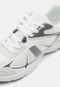 Valkoinen urheilushoe, jossa on verkkokangasta, synteettisiä yksityiskohtia ja harmaata sävyä. Sisältää kontrastivärisiä mustia viivoja ja pyöreitä naruja.