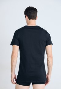 Man seen from the back wearing a plain black short-sleeve t-shirt and black underwear against a neutral light background.