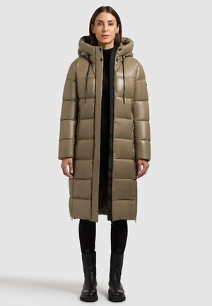 Geacă puffer de culoare măslinie, cu glugă și fermoar negru, având un design matlasat, mâneci lungi și fermoare laterale. Puritată deasupra unor pantaloni negri și cizme.