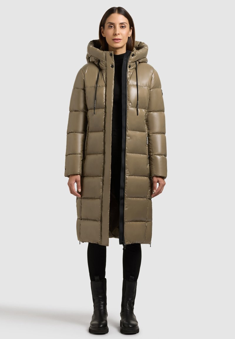 Olivová pufová bunda s kapucňou a čiernym zippers, s prešívaným dizajnom, dlhými rukávmi a bočnými zipsami. Nosená cez čierne nohavice a topánky.