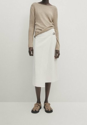Femme portant un pull à manches longues beige rentré dans une jupe midi blanche avec une boucle métallique, assorti à des sandales beiges fermées.