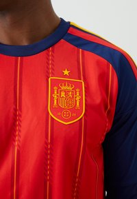 Röd och blå sporttröja med spansk landslagsmärke kantat i gult, som visar en krona, sköld, stjärna och emblemet "RFEF 1909".