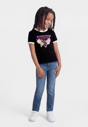 Ragazza con capelli intrecciati che indossa una T-shirt nera a fiori Guess, jeans blu e sneakers bianche, in piedi e sorridente su uno sfondo bianco.