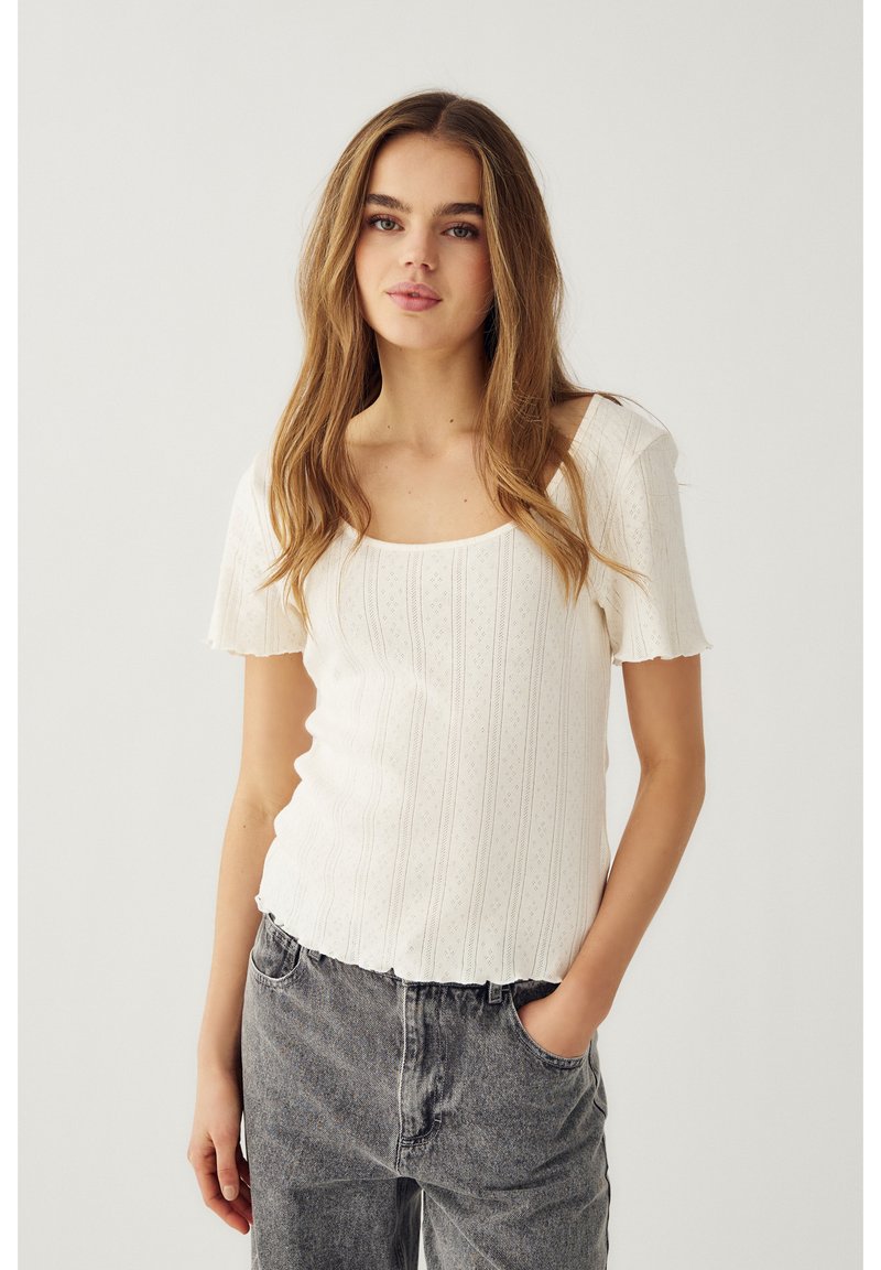 Junge Frau mit langen hellbraunen Haaren, die ein weißes, strukturiertes Stricktop mit kurzen Ärmeln und graue, hoch taillierte Jeans trägt, eine Hand in der Tasche.