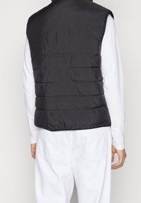 Schwarze Steppweste mit gestepptem Design, hohem Kragen und Reißverschluss, kombiniert mit einem weißen Langarmshirt und hellen Hosen.