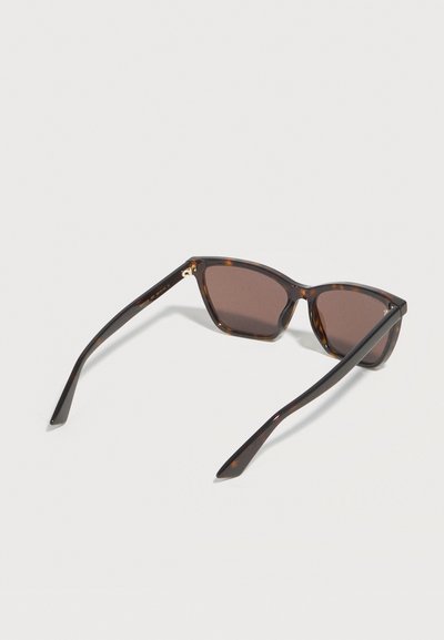 Γυαλιά ηλίου με καφέ ταρταρούγα σκελετό, σκουρόχρωμους ορθογώνιους φακούς και λεπτούς καμπύλους βραχίονες, ανοιχτά πάνω σε ανοιχτό γκρι επιφάνεια.