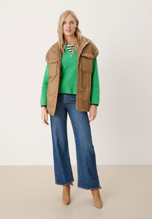 Gilet rembourré marron avec poches à rabat sur un pull en maille vert, associé à un jean bleu à jambes larges et des escarpins beiges fermés.